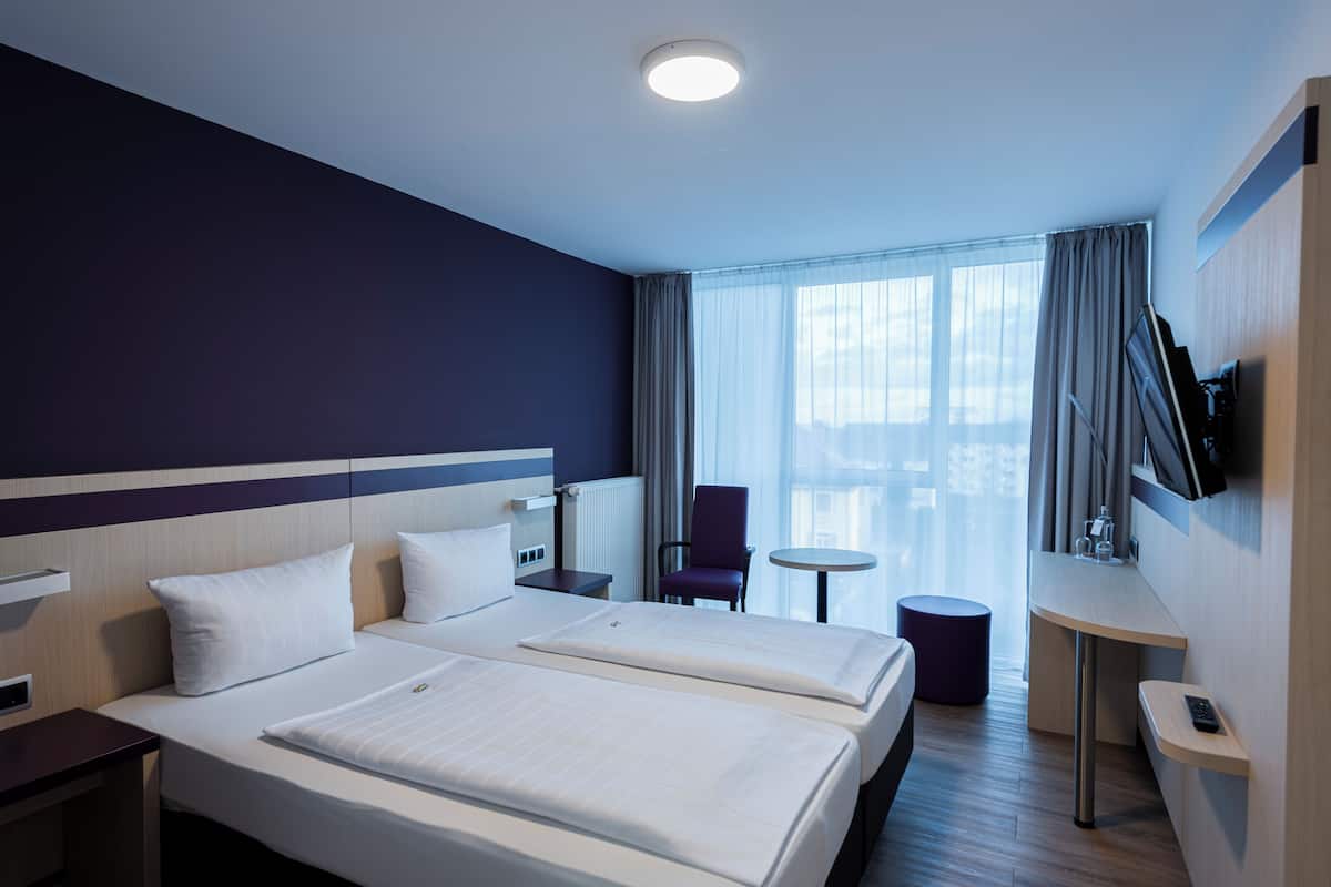 A hotel room with two beds, a small table, a chair, a TV mounted on the wall, and a window with curtains.