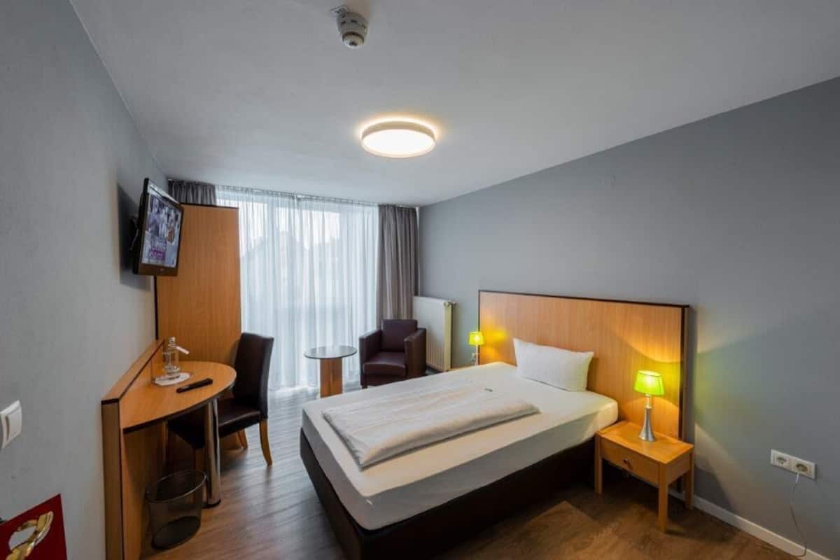 A hotel room with a bed, bedside tables, a desk, a chair, a television, and a window with curtains.