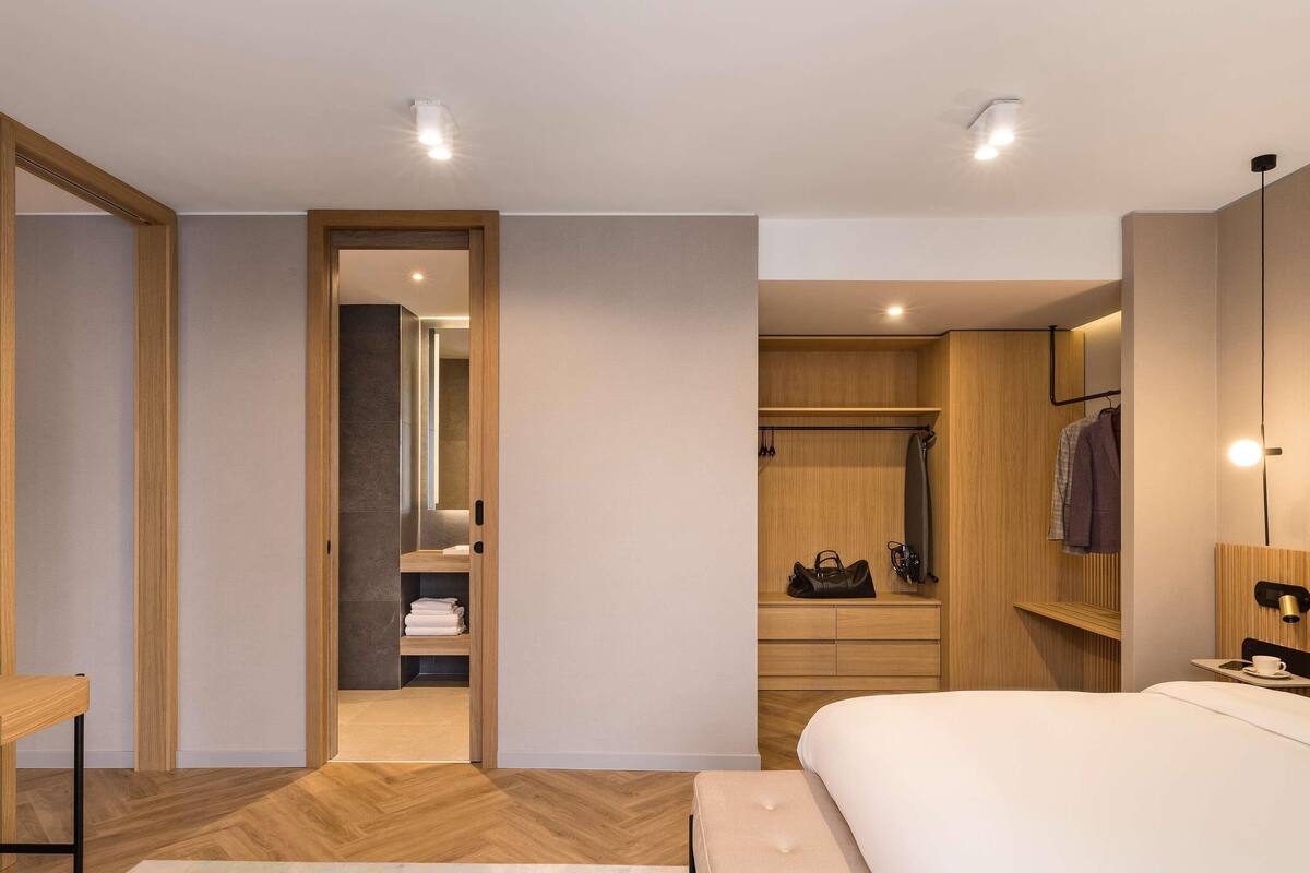 A modern hotel room with a wooden floor, a large bed, a bedside table, a wardrobe, and a bathroom visible through an open door.