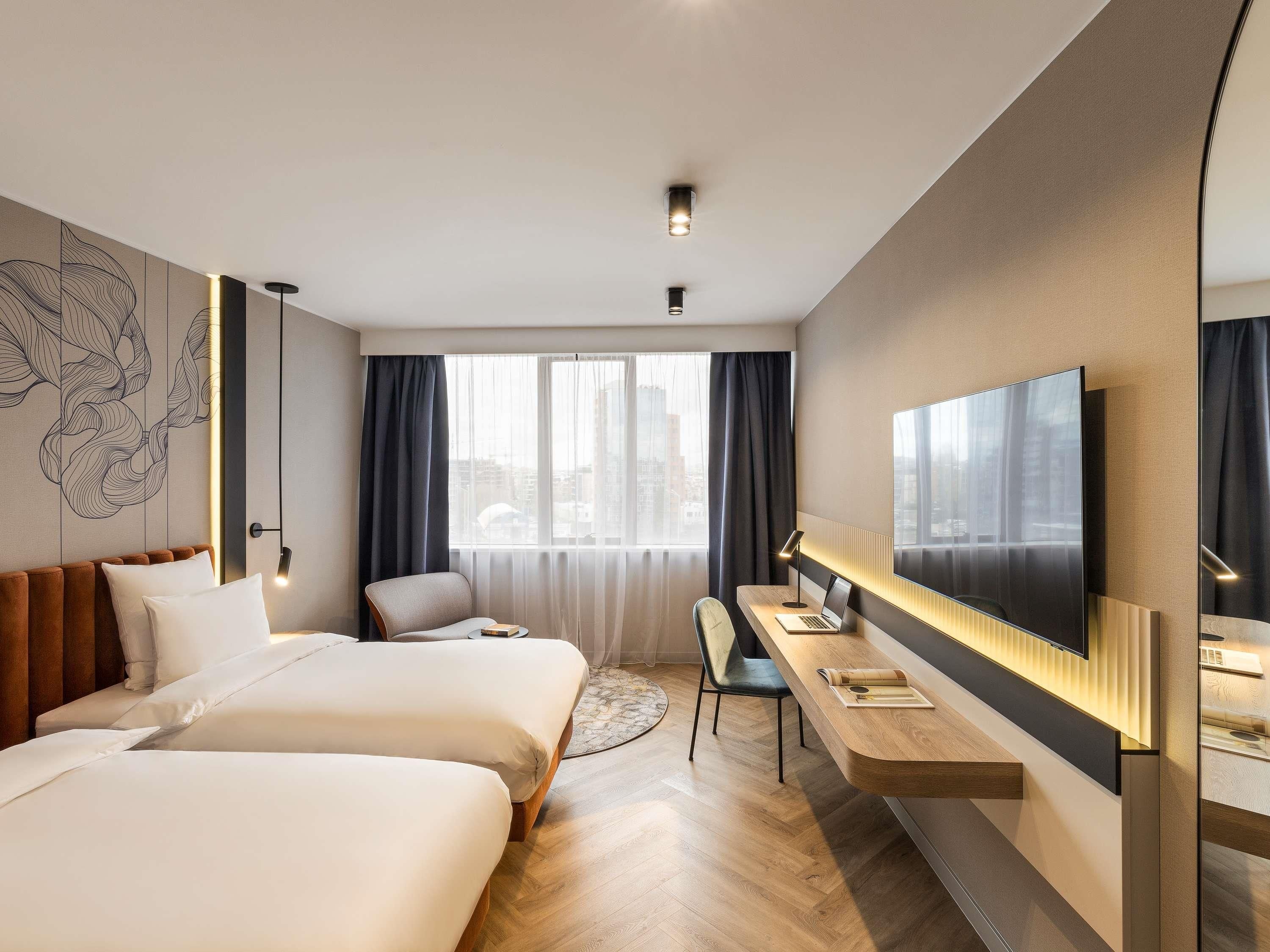 A modern hotel room with two beds, a desk, a chair, and a large TV.