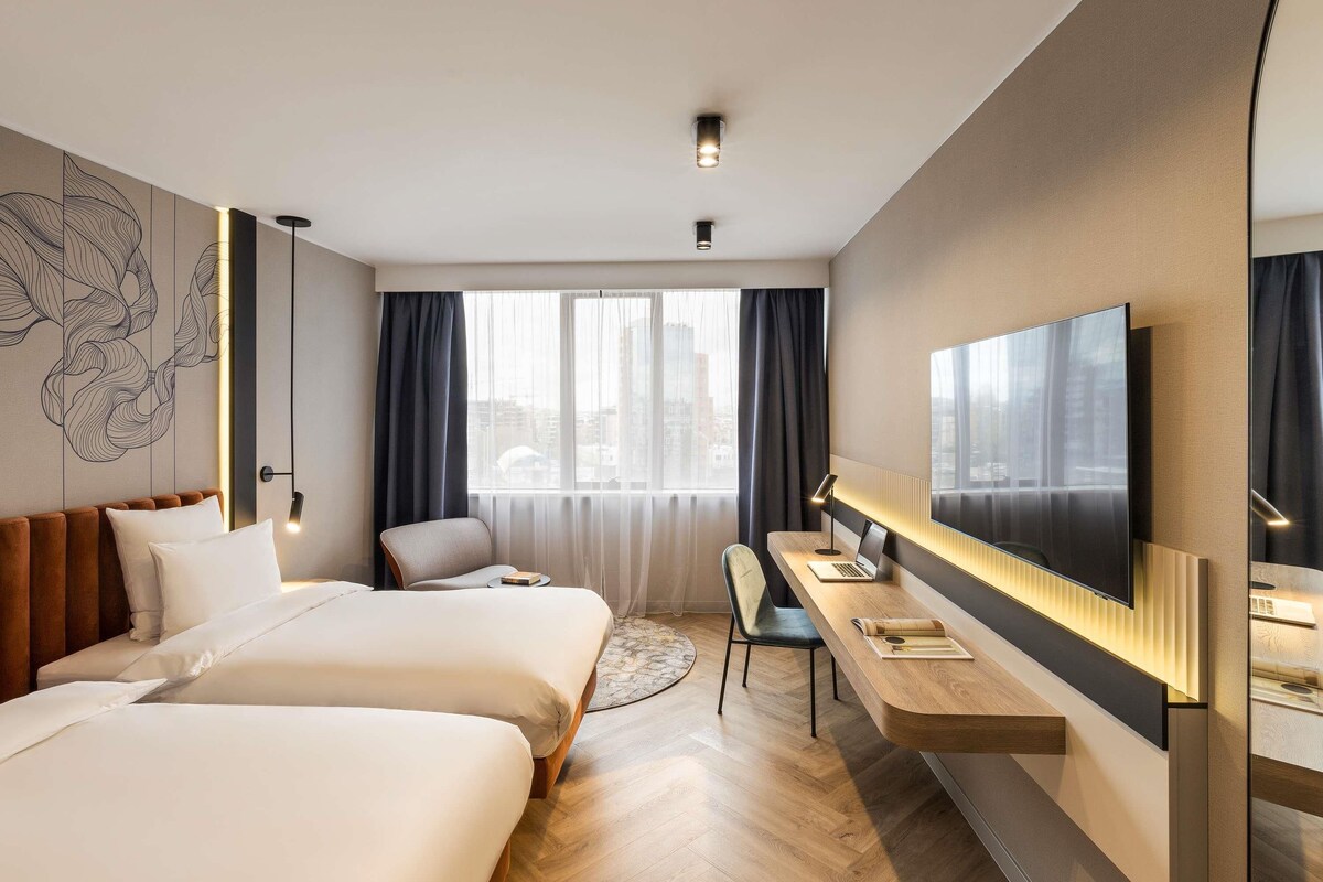 A modern hotel room with two beds, a desk, a chair, and a large TV.