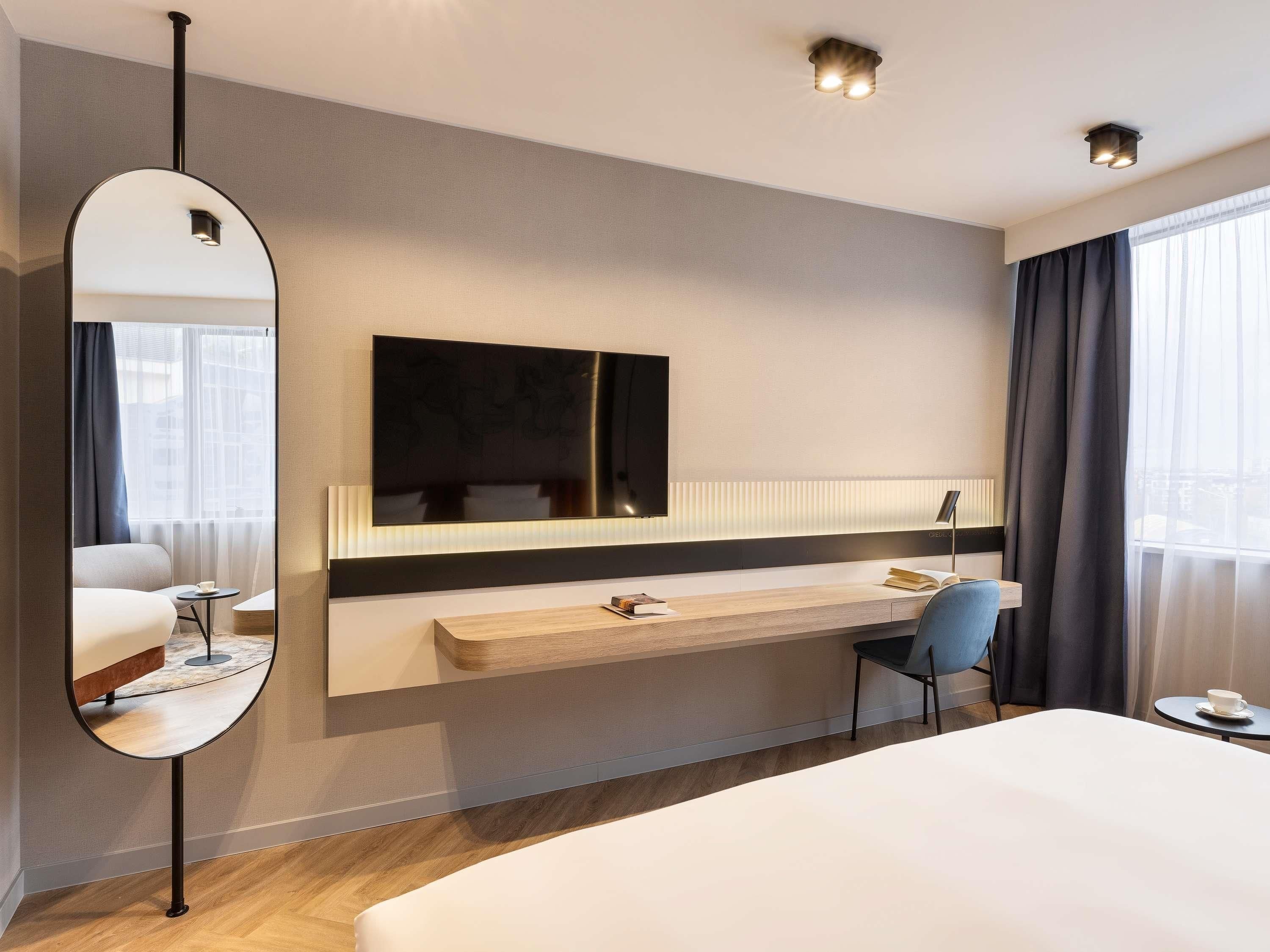 A modern hotel room with a large bed, a bedside table, a desk, a chair, a large mirror, and a flat-screen TV mounted on the wall.