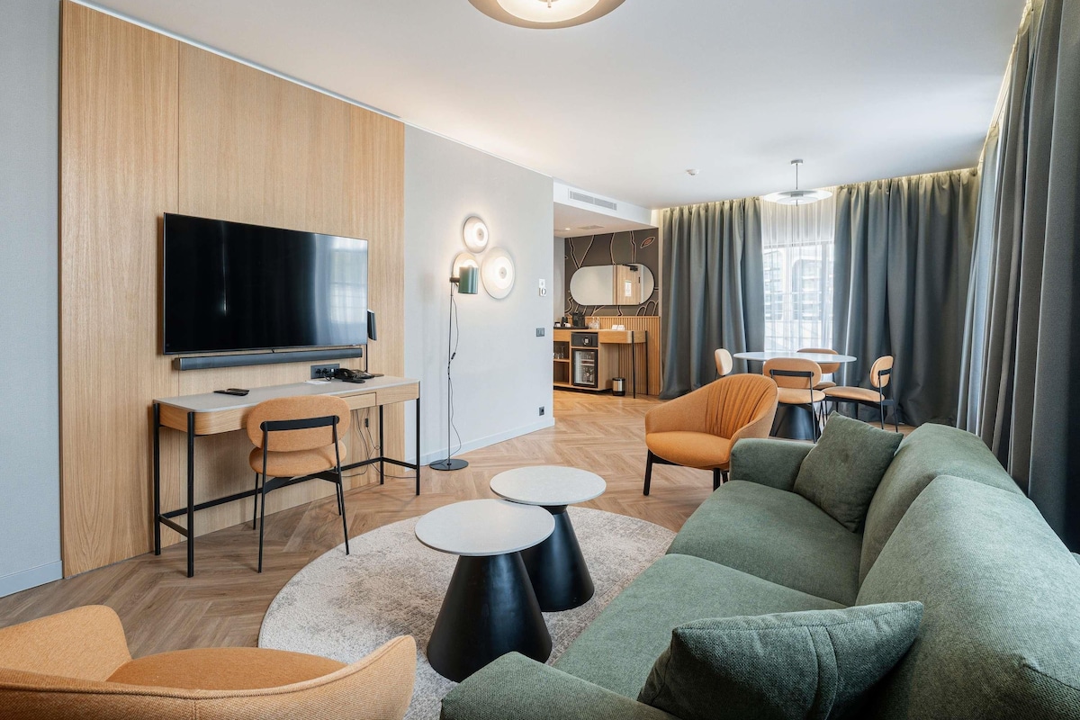 A modern living room with a flat-screen TV, a sofa, two round tables, and a dining area in the background.