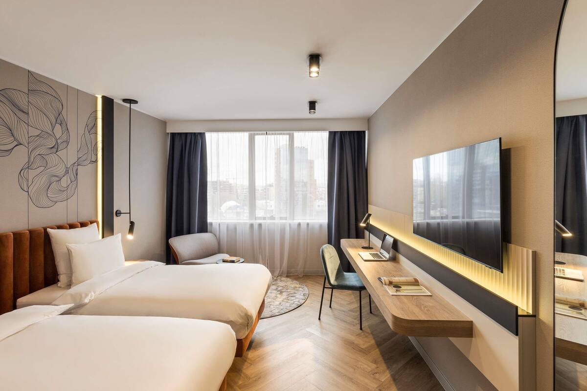 A modern hotel room with two beds, a desk, a chair, and a large TV.