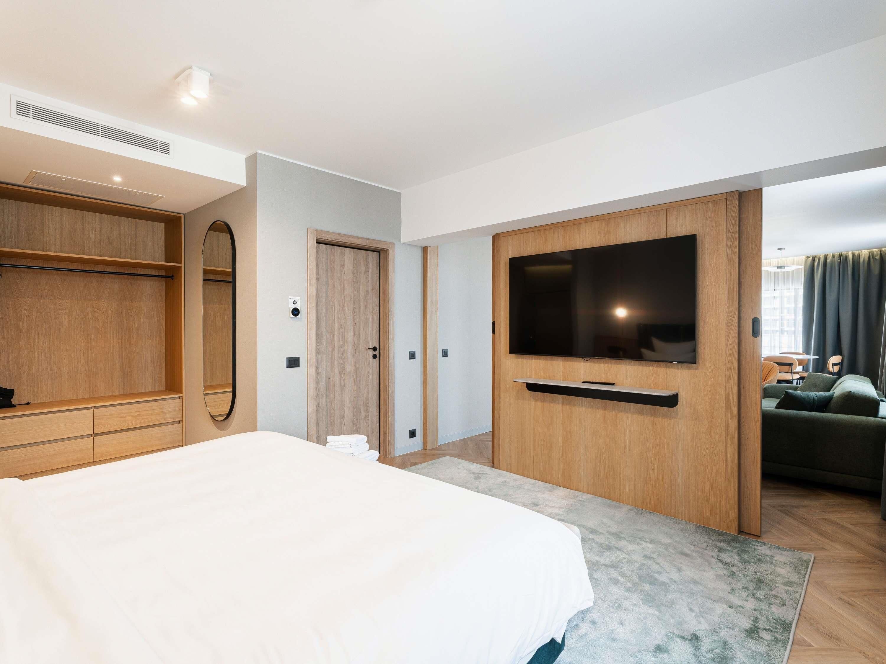 A modern hotel room with a large bed, a TV mounted on a wooden panel, a wardrobe, and a view of a dining area.