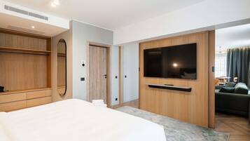 A modern hotel room with a large bed, a TV mounted on a wooden panel, a wardrobe, and a view of a dining area.