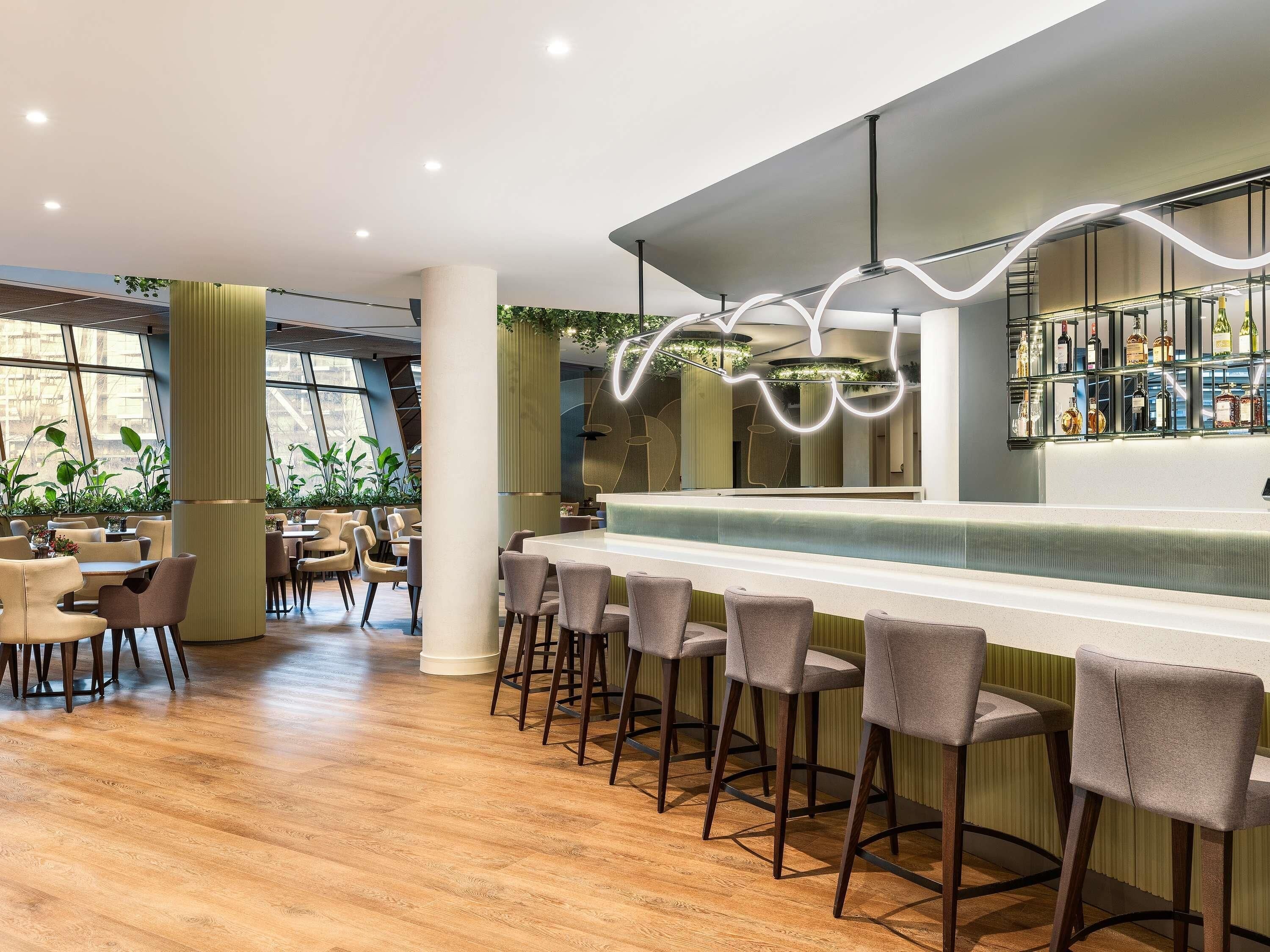 A modern bar area with a long counter and stools, surrounded by tables and chairs, with large windows and indoor plants.