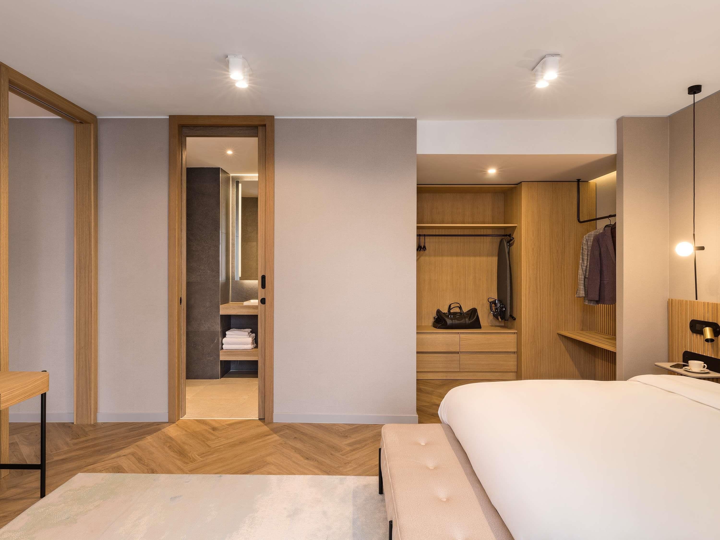 A modern hotel room with a wooden floor, a large bed, a bedside table, a wardrobe, and a bathroom visible through an open door.