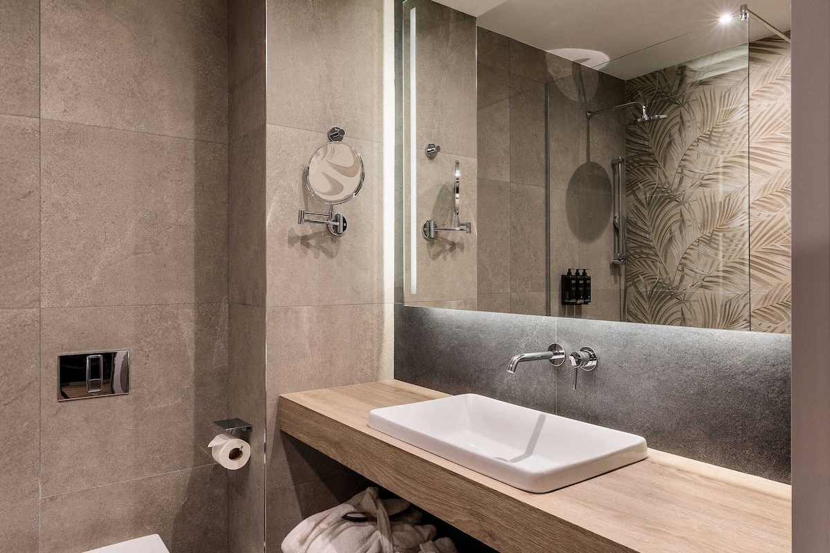 A modern bathroom with a large mirror, a sink, and a toilet. There are towels and toiletries on the vanity.