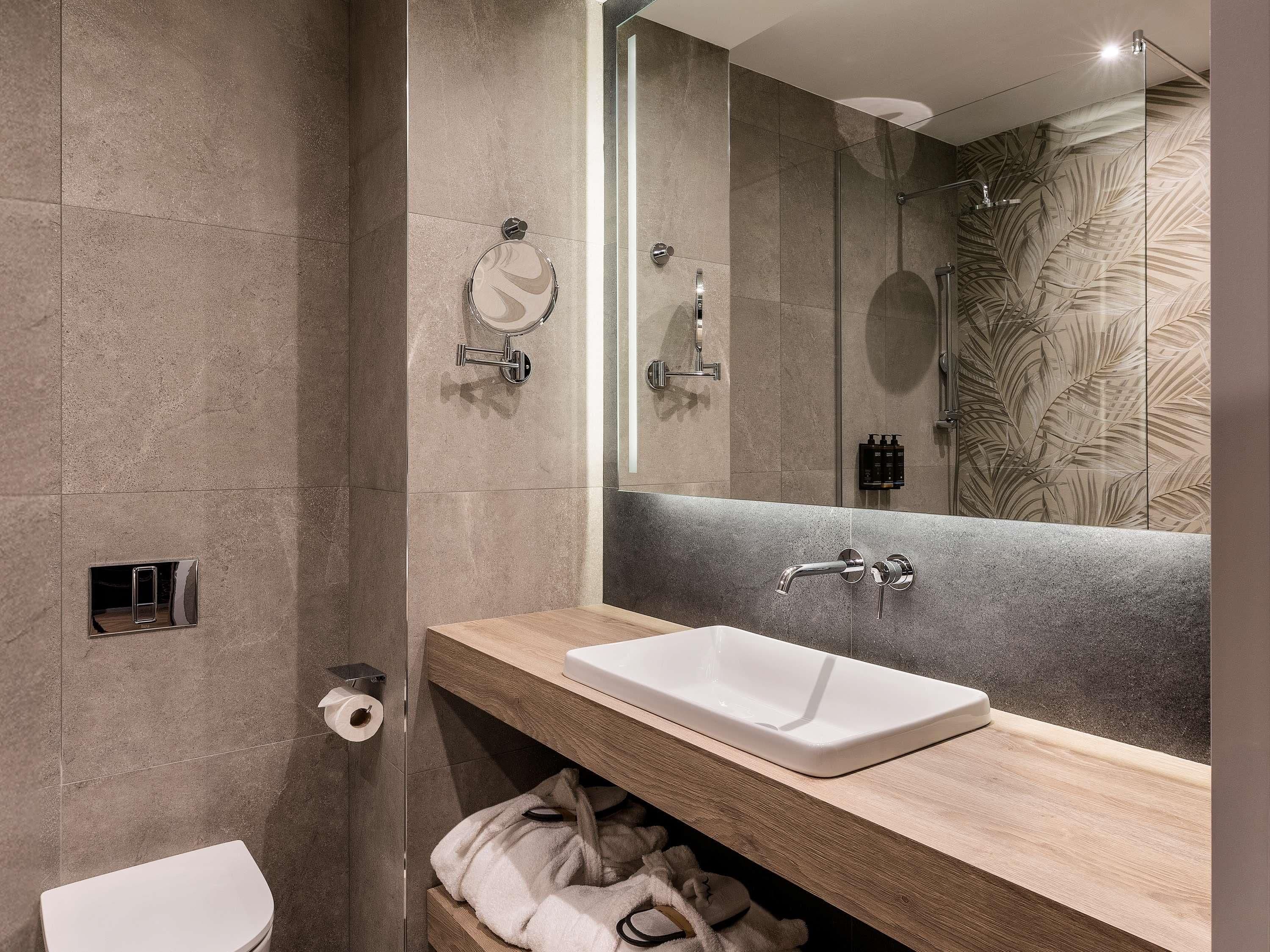 A modern bathroom with a large mirror, a white sink, a shower, and a towel rack.