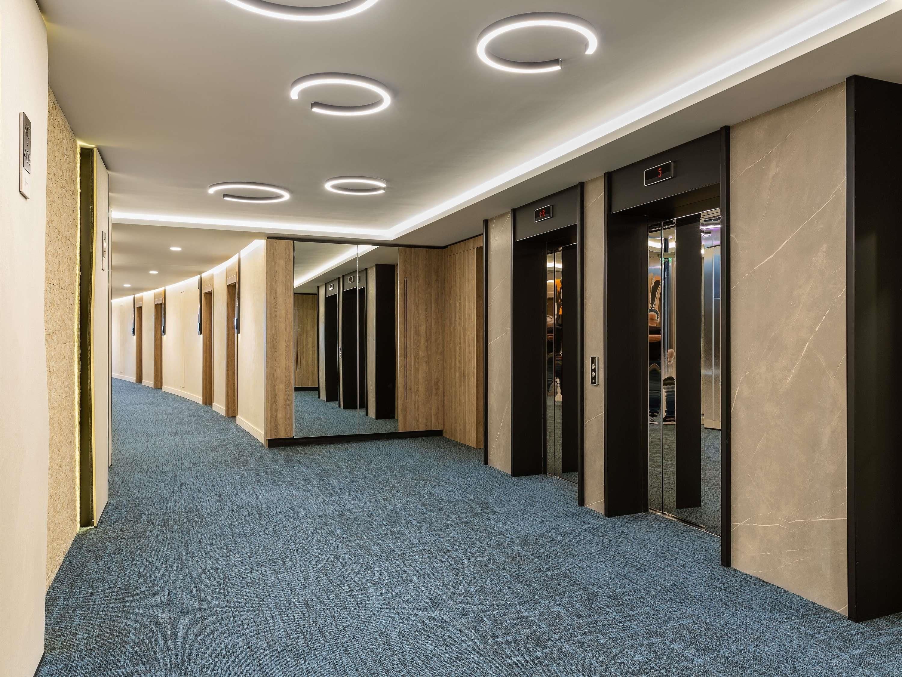 Un couloir d’hôtel avec une moquette bleue, des murs en bois et des luminaires modernes.