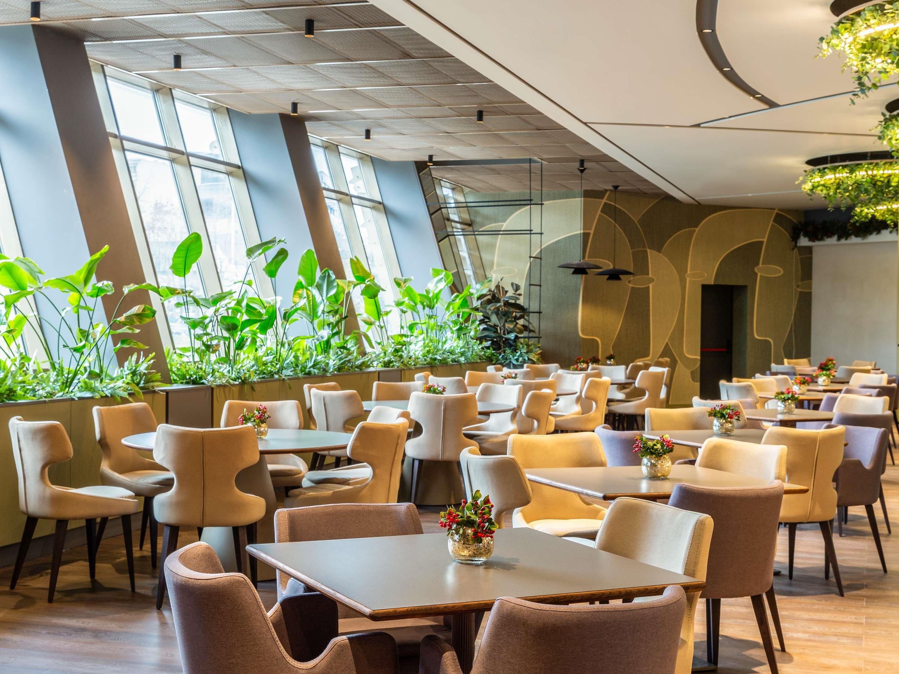 A modern restaurant interior with high ceilings, large windows, and indoor plants.