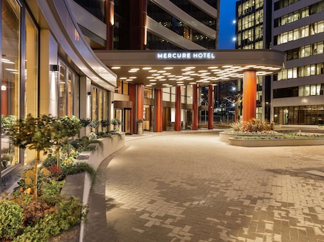 The exterior of the Mercure Hotel with a modern design, featuring a prominent entrance with a canopy and a landscaped area with plants.. Mercure Sofia City