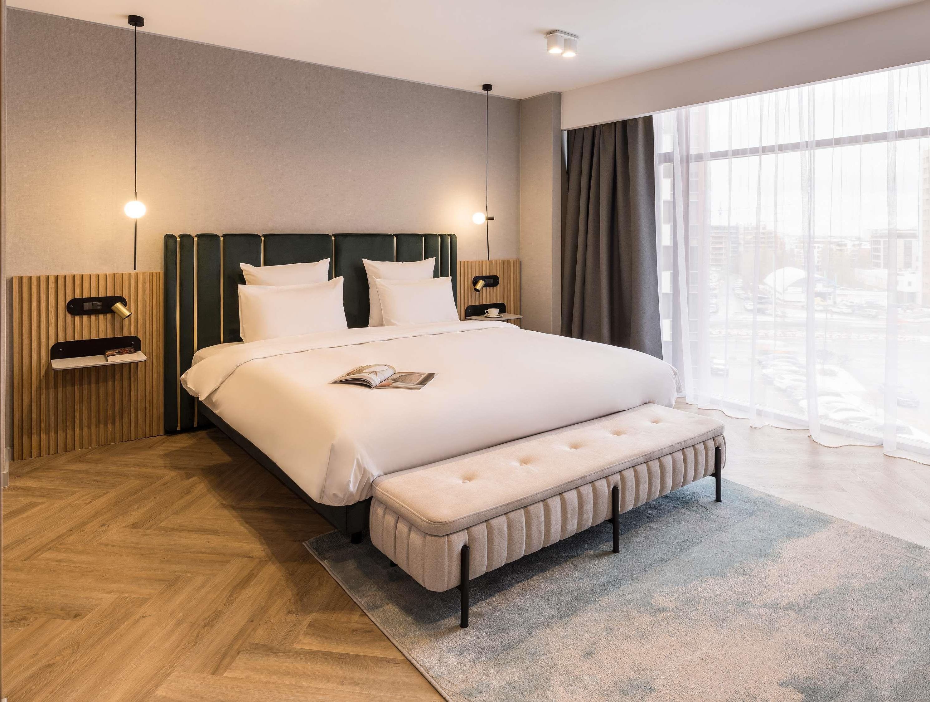 A modern hotel room with a large bed, a bench, a bedside table, and a view of the city through large windows.