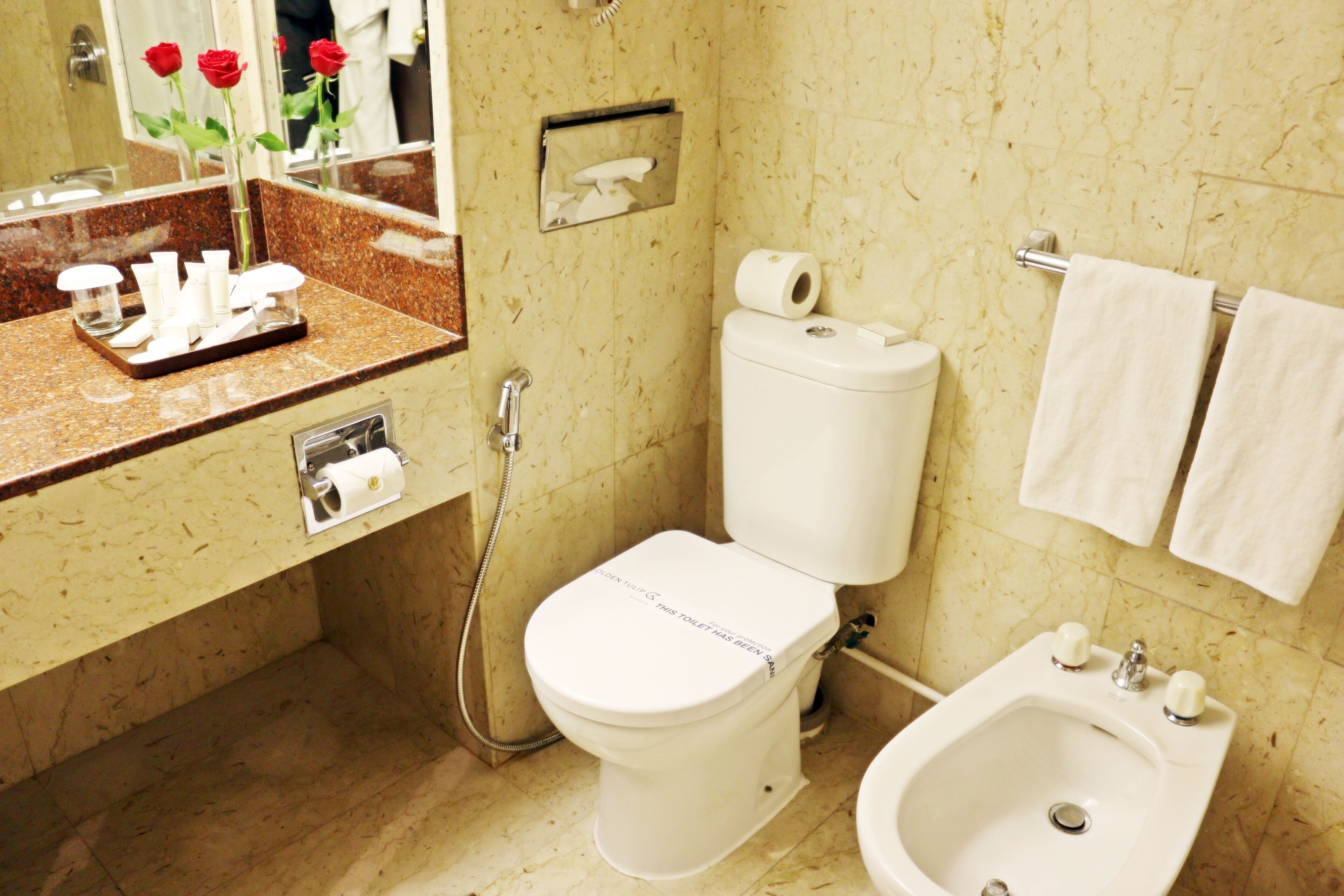 A bathroom with a toilet, bidet, sink, and a mirror with a rose vase.