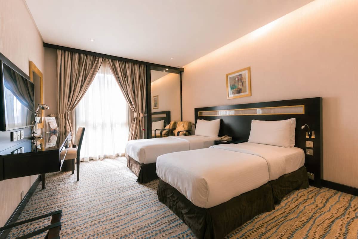 A hotel room with two beds, a desk, a chair, a TV, and a large window with curtains.