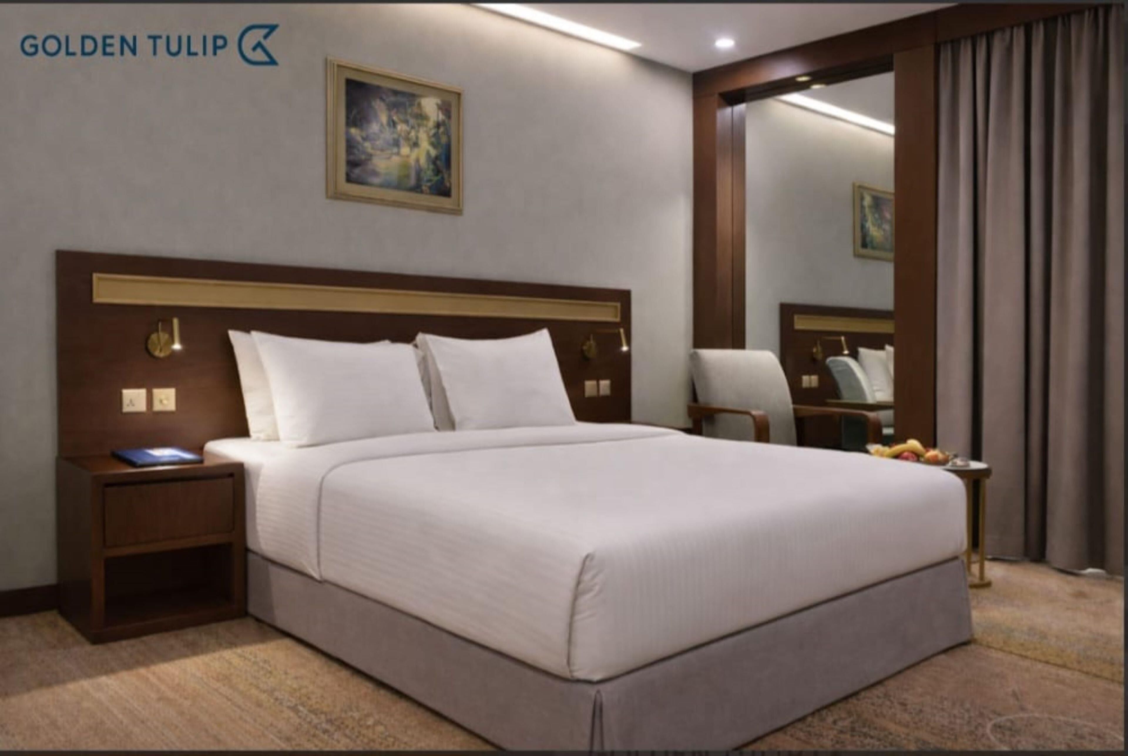 A hotel room with a large bed, a nightstand with a lamp, a chair, a mirror, and a painting on the wall.