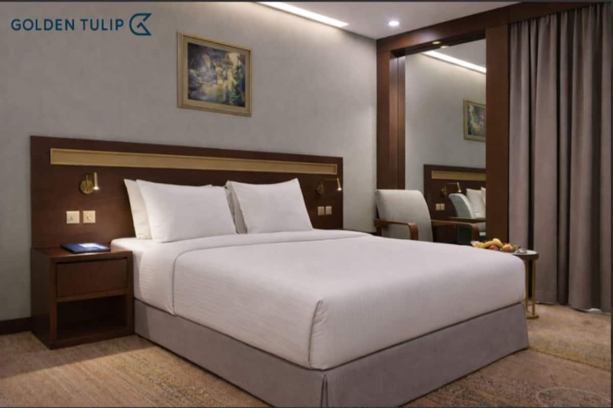 A hotel room with a large bed, a nightstand with a lamp, a chair, a mirror, and a painting on the wall.