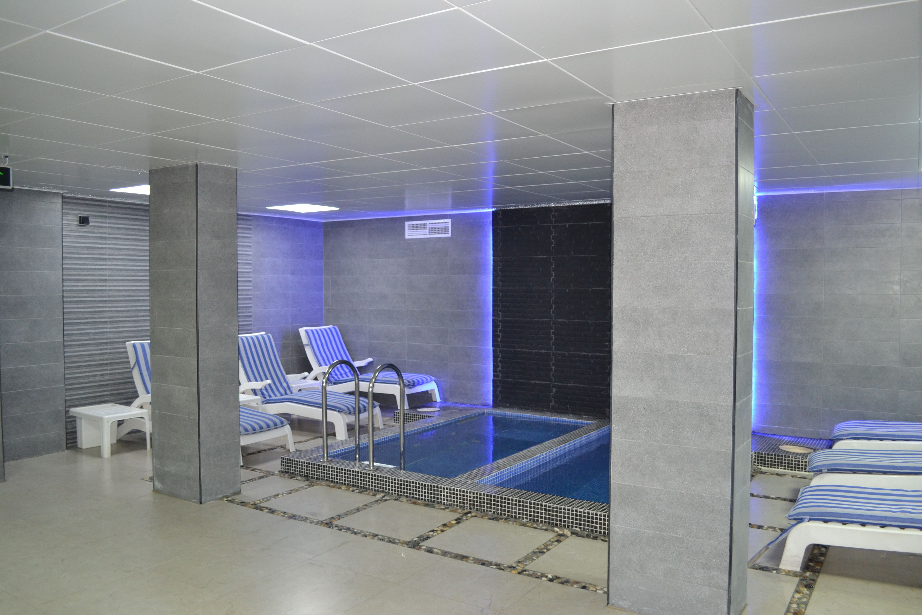 A modern indoor swimming pool with blue and white striped lounge chairs, a small poolside table, and a tiled floor.