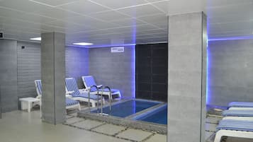 A modern indoor swimming pool with blue and white striped lounge chairs, a small poolside table, and a tiled floor.