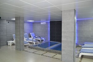 Una piscina interior moderna con sillas de salón azul y blanco a rayas, una pequeña mesa junto a la piscina y suelo de baldosas.