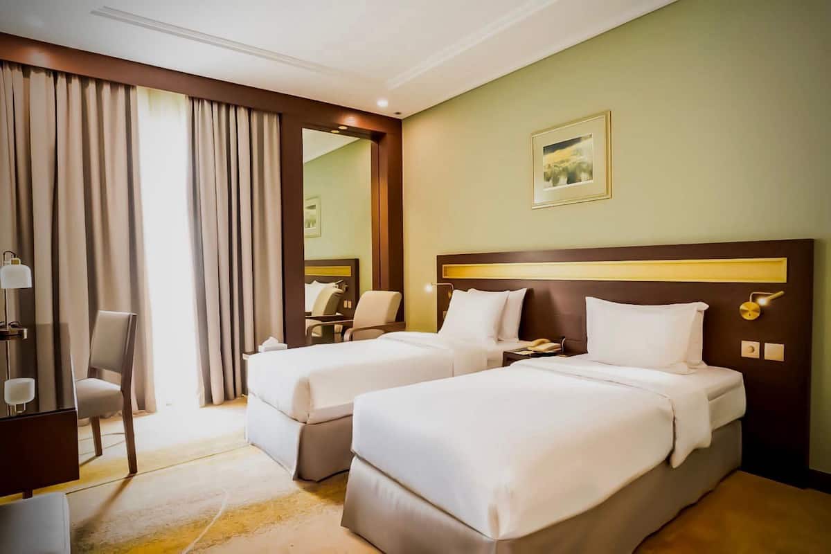A hotel room with two beds, a desk, a chair, and a large window with curtains.