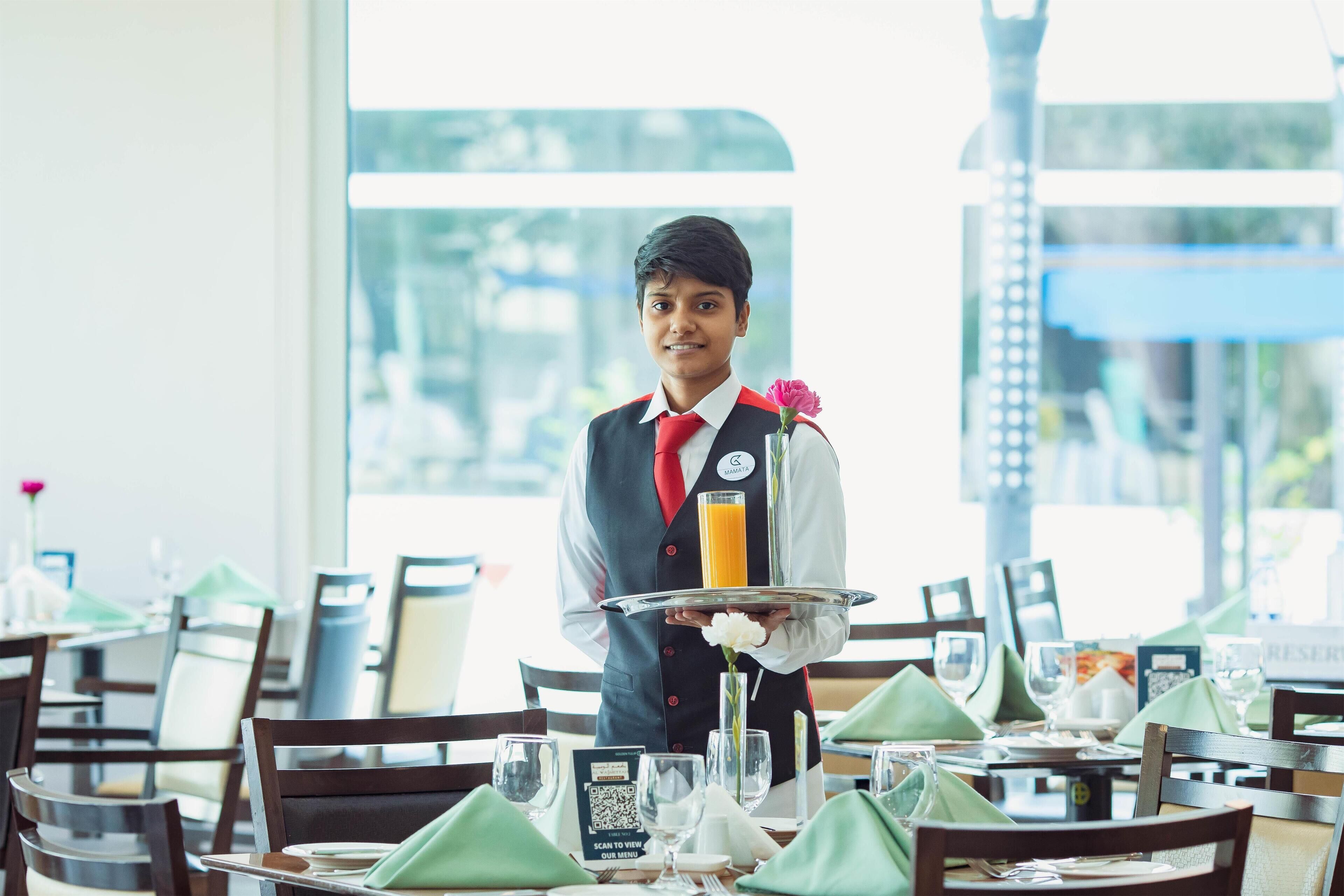 A waiter in a restaurant holding a tray with a drink and a flower. Tables are set with green napkins and silverware.