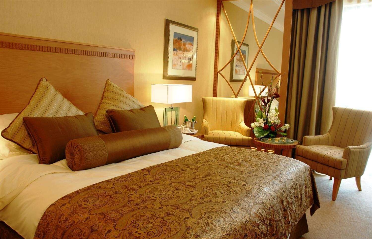 A hotel room with a large bed, multiple pillows, a chair, a small table with a vase, and a lamp.