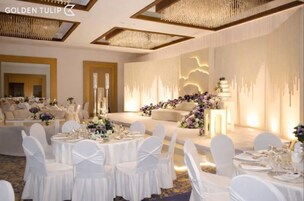Un salón de banquetes con mesas redondas, sillas blancas y un escenario decorado con arreglos florales.