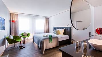 Una moderna camera d'albergo con un ampio letto, un'area salotto con una poltrona verde, un bagno con un grande specchio e lavandino, e vista sulla città.