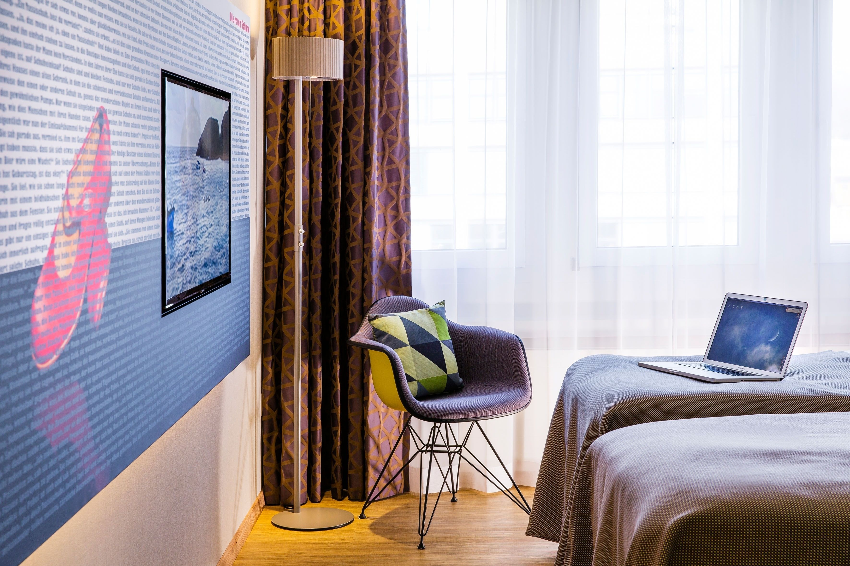 Camera d'albergo con un letto, una poltrona con un cuscino colorato, un laptop sul letto, un'ampia finestra e una parete con un'immagine e del testo.