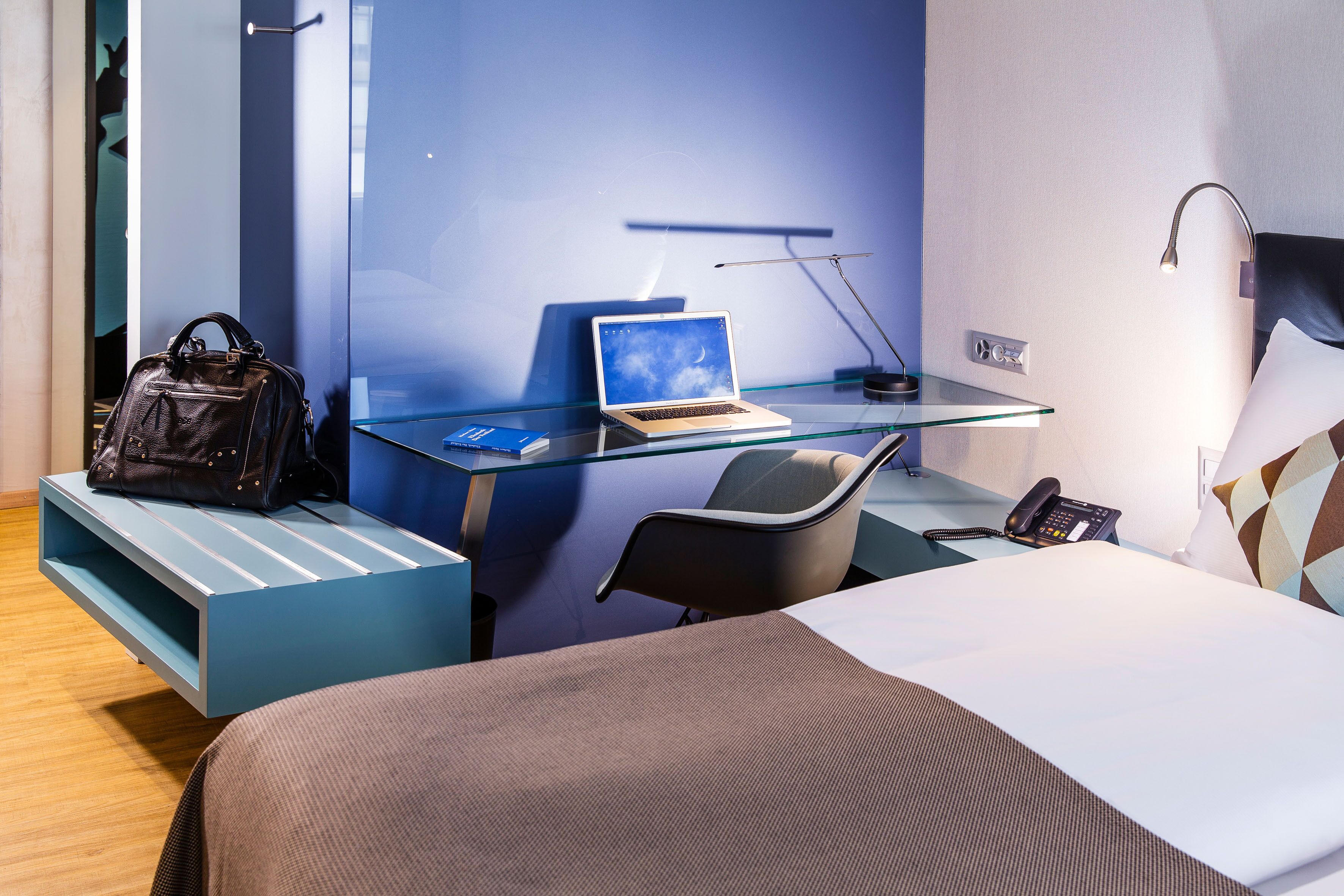 A hotel room with a bed, a desk with a laptop, a chair, a desk lamp, a phone, and a handbag on a bench.