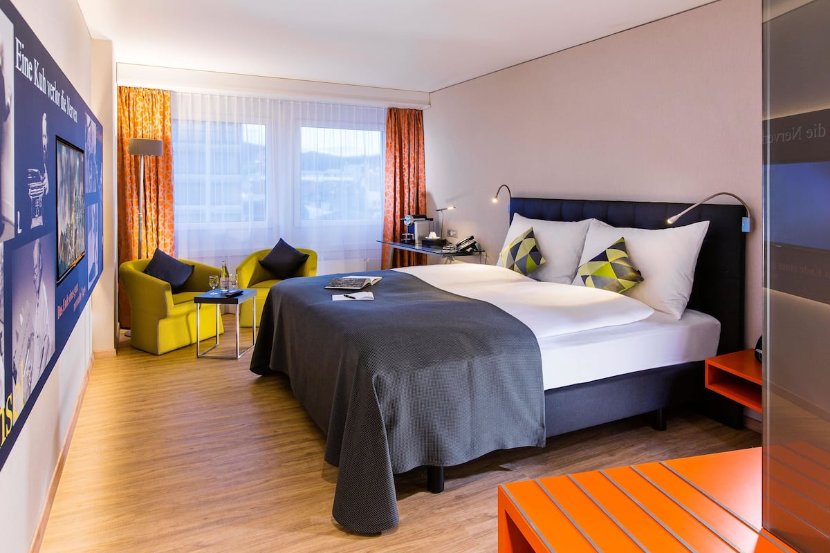 A modern hotel room with a large bed, a desk, a chair, and a sofa.