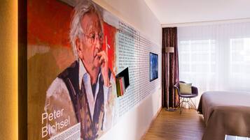 A hotel room with a large portrait on the wall, a bed, a chair, and a window with curtains.
