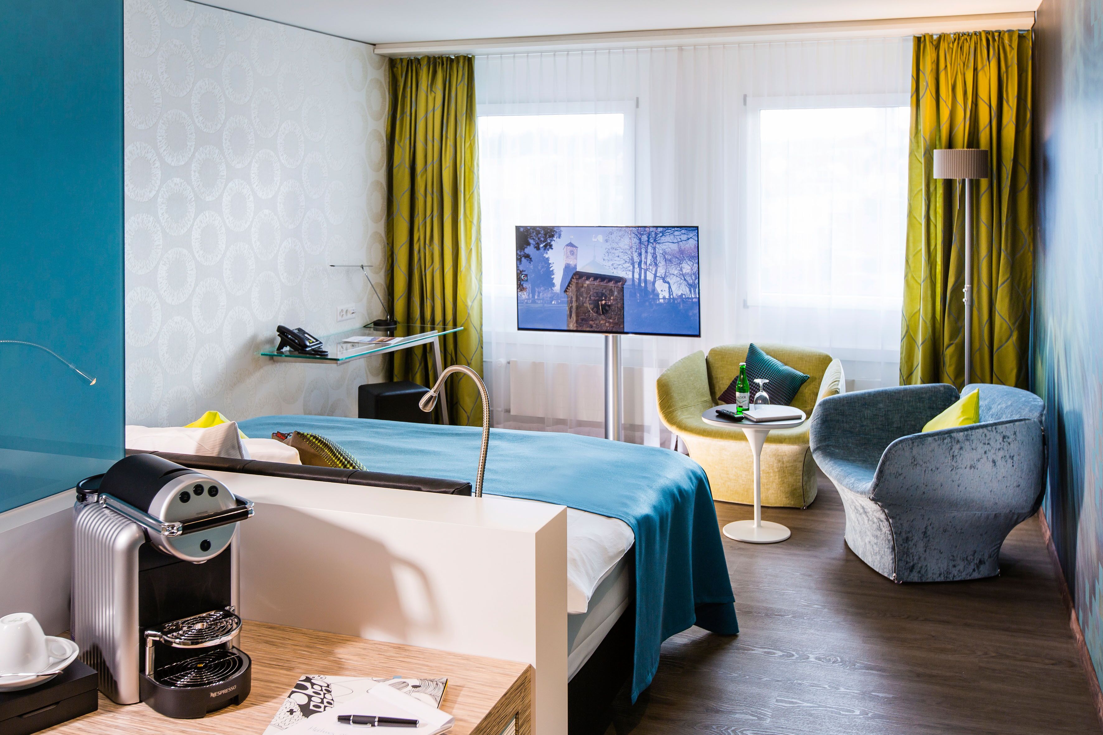 Habitación de hotel con cama, escritorio con cafetera, televisor, silla y lámpara.