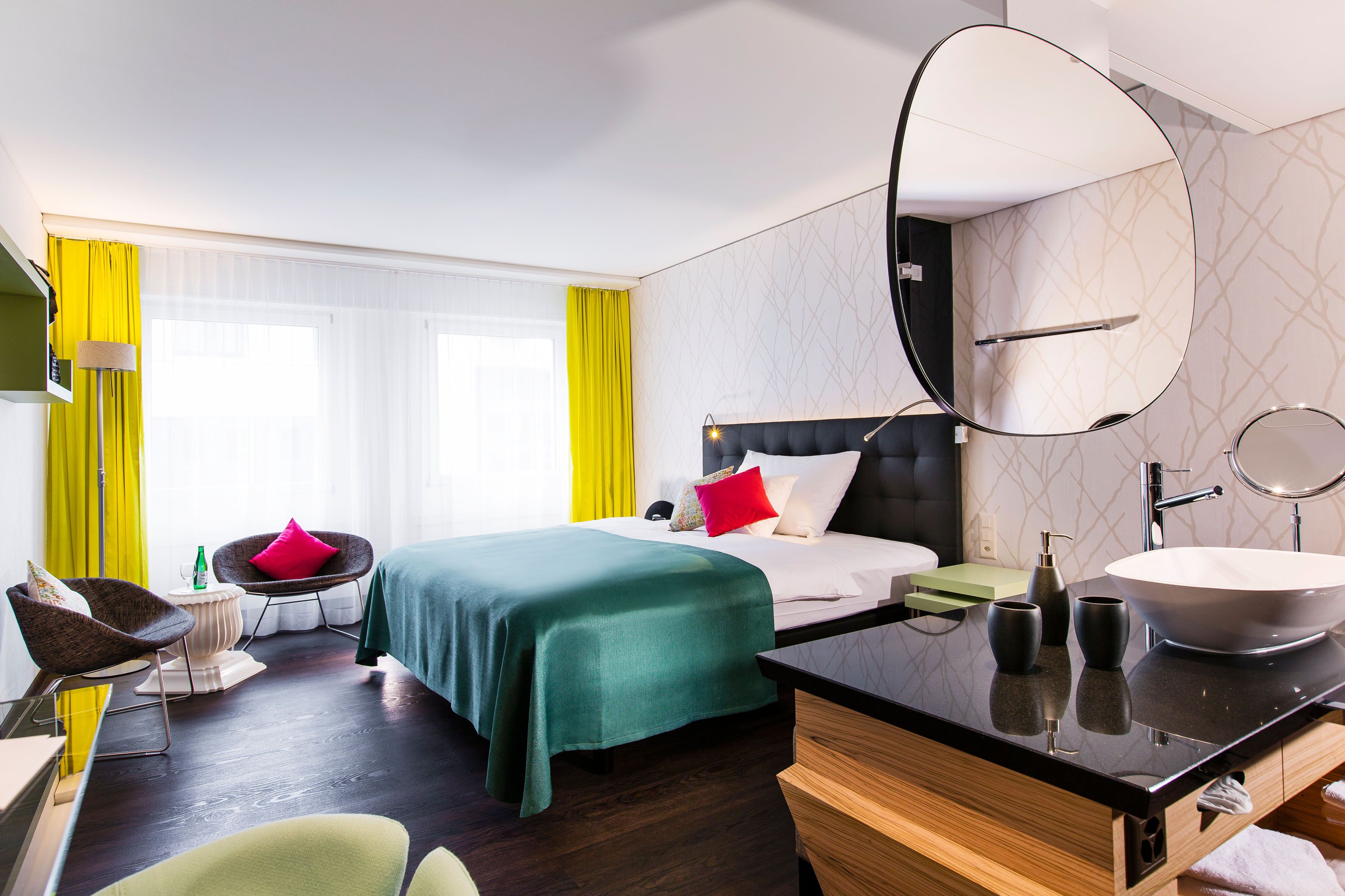 A hotel room with a bed, a desk, a chair, a large mirror, a sink, and a window with yellow curtains.