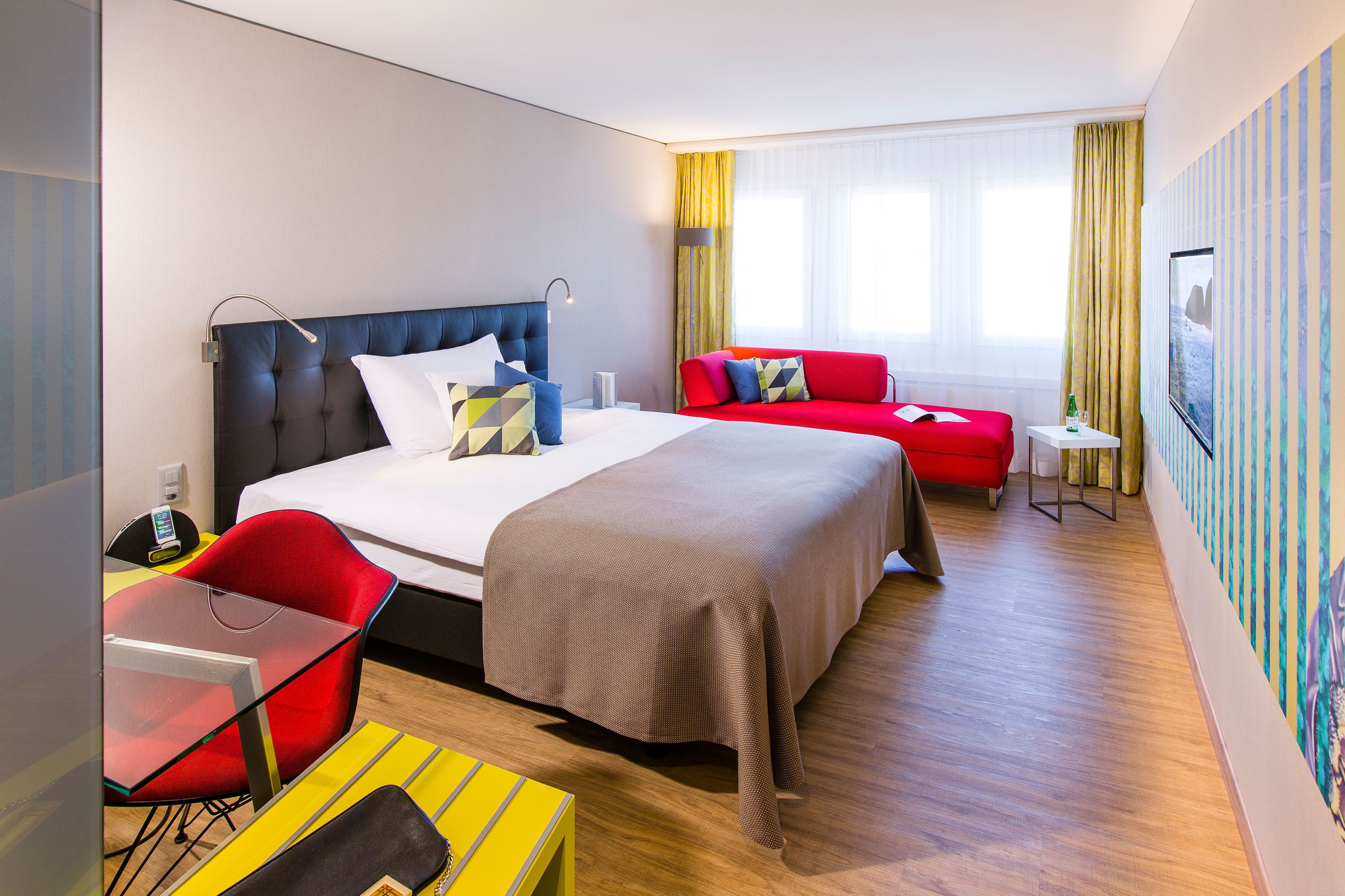 Ein modernes Hotelzimmer mit einem großen Bett, einem roten Sofa, einem Glastisch und einem kleinen Nachttisch.