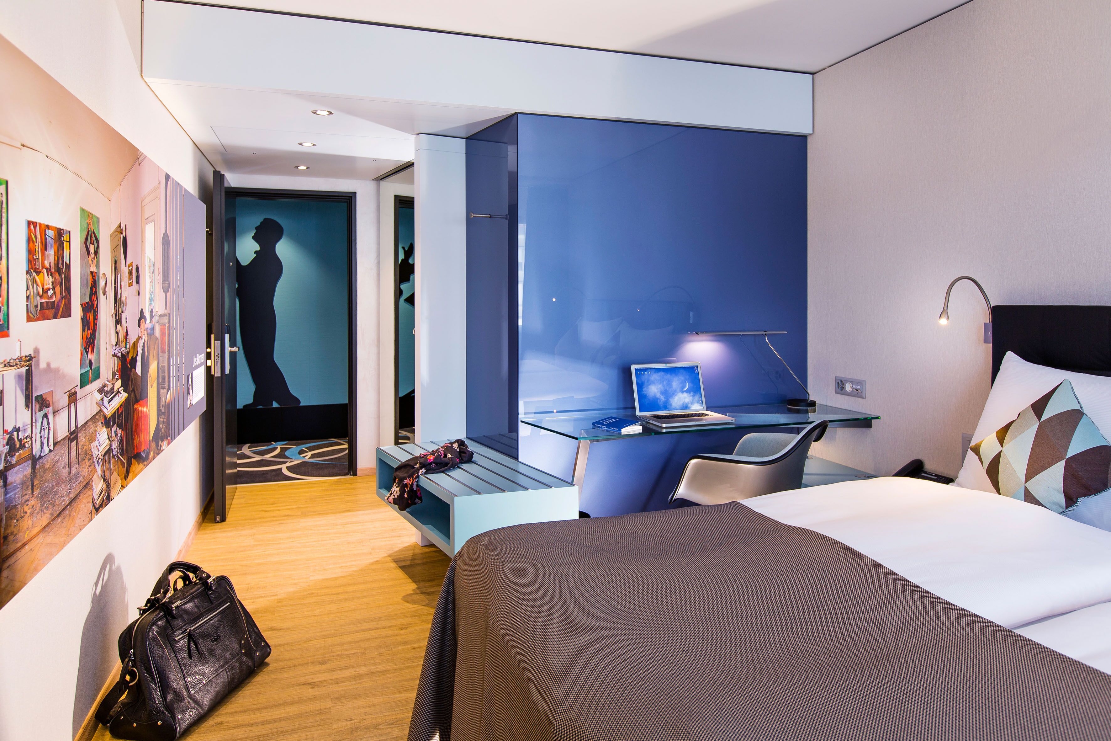 Una habitación de hotel moderna con cama, escritorio y zona de estar. Hay un ordenador portátil sobre el escritorio y un bolso en el suelo.