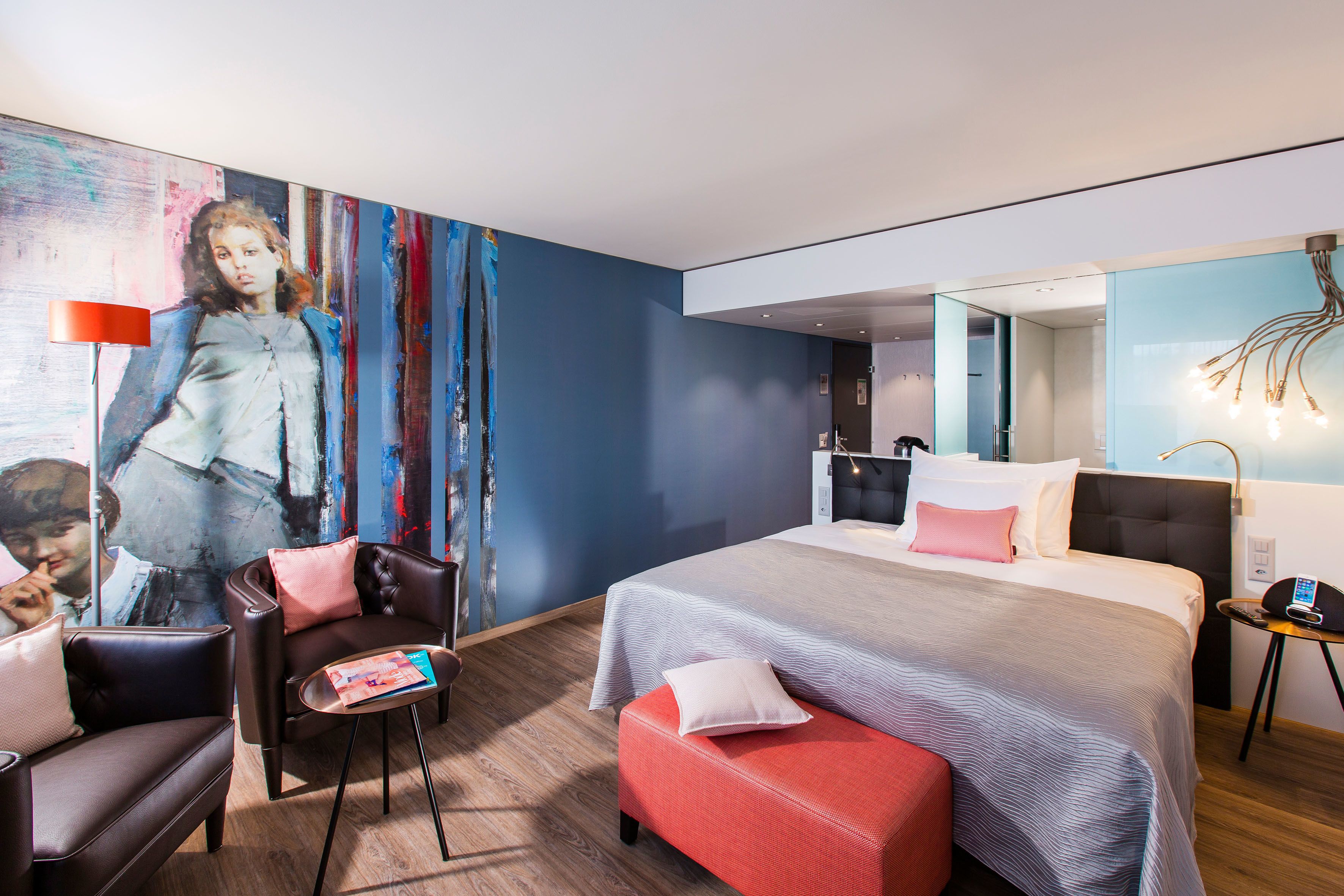 Een moderne hotelkamer met een groot bed, een zithoek met een rode poef, een bijzettafeltje en een schilderij aan de muur.
