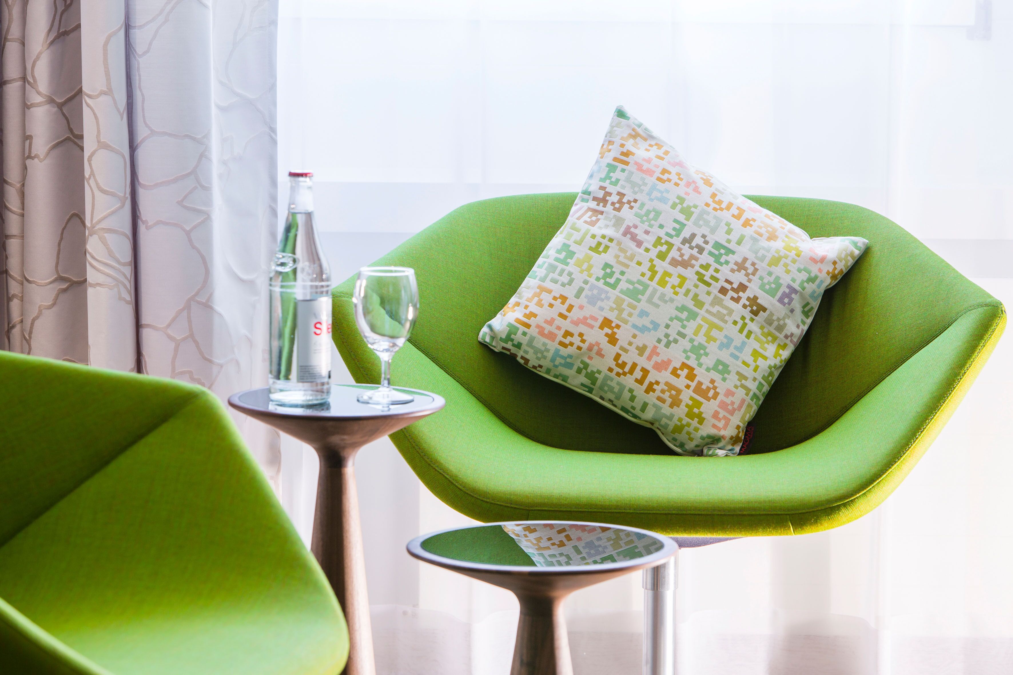 Une pièce avec un fauteuil vert, une petite table ronde sur laquelle se trouvent une bouteille et un verre, et un coussin aux couleurs vives.