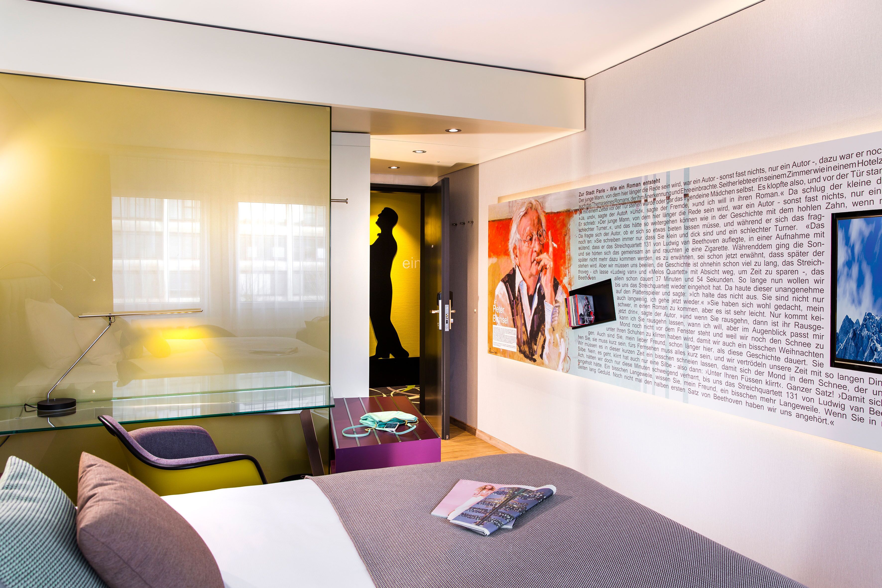 Une chambre d’hôtel moderne avec un bureau en verre, un lit avec un magazine et un mur orné d’un texte et d’une affiche.