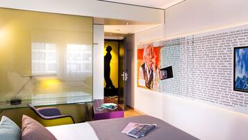 Une chambre d’hôtel moderne avec un bureau en verre, un lit avec un magazine et un mur orné d’un texte et d’une affiche.