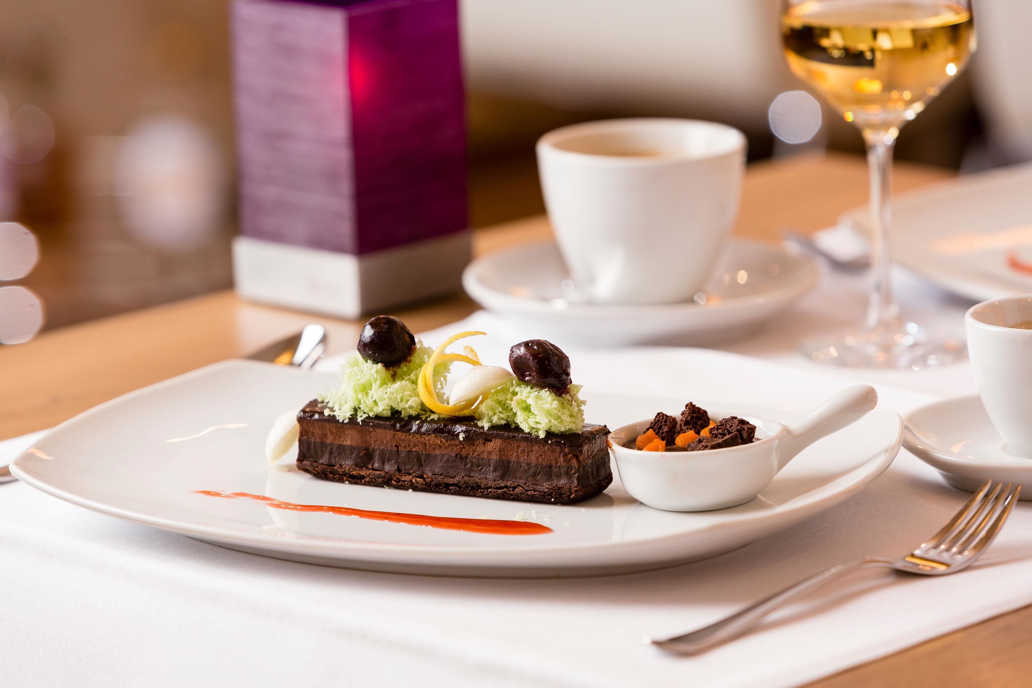 A dessert plate with a chocolate cake, a small bowl of sauce, and a glass of white wine on a table.