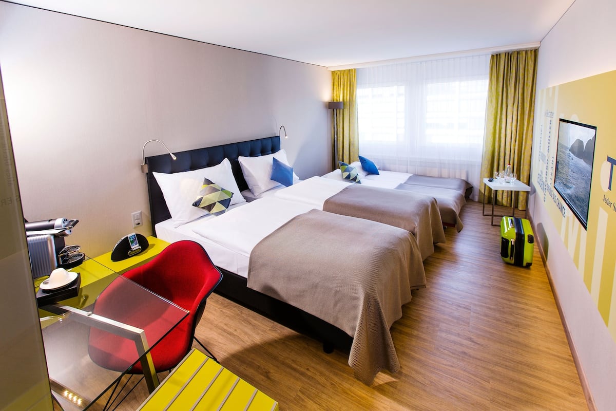 A hotel room with two beds, a desk with a chair, a small table with a lamp, and a suitcase on the floor.