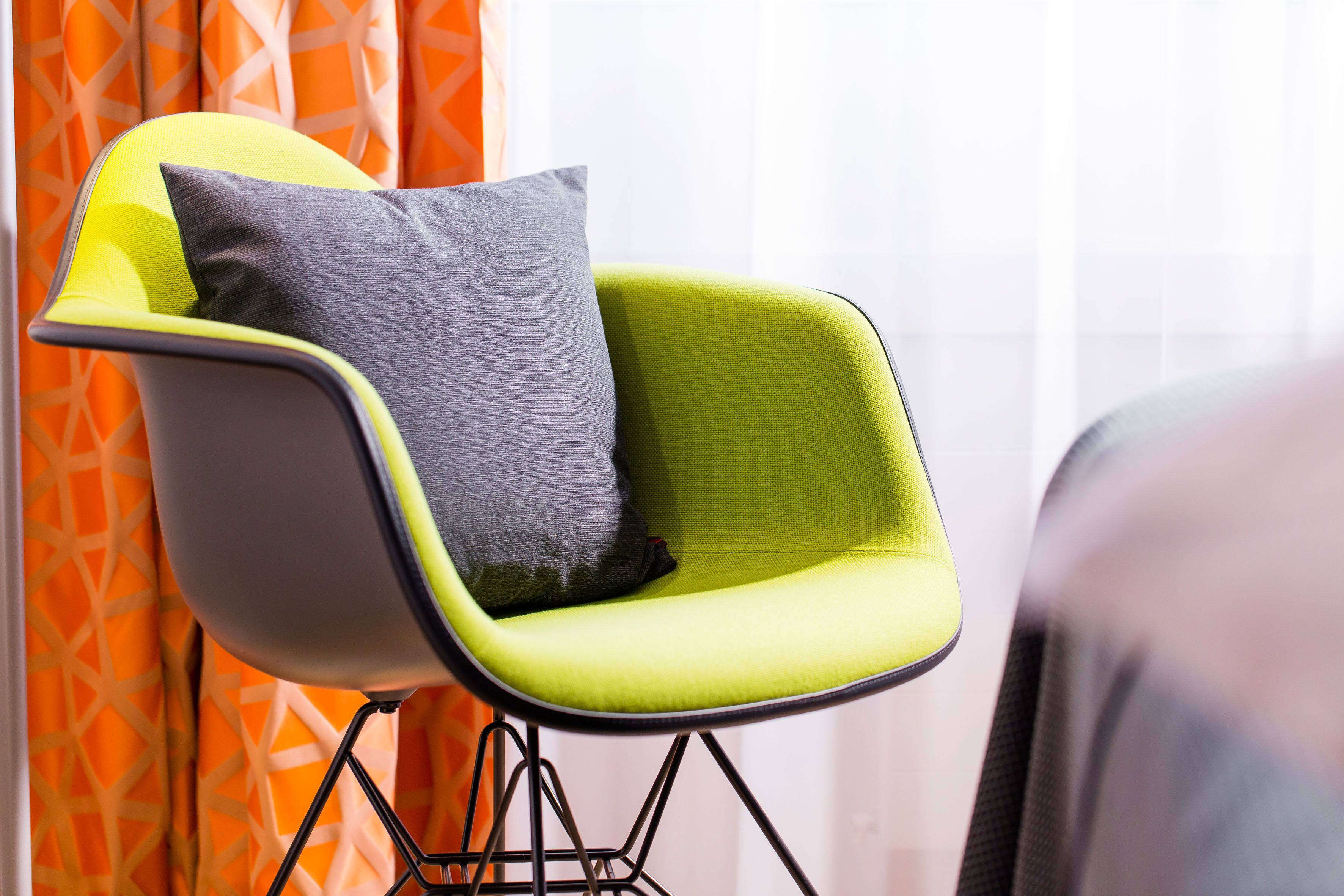 Una silla moderna con asiento verde y cojín gris, ubicada frente a una ventana con cortinas naranjas.