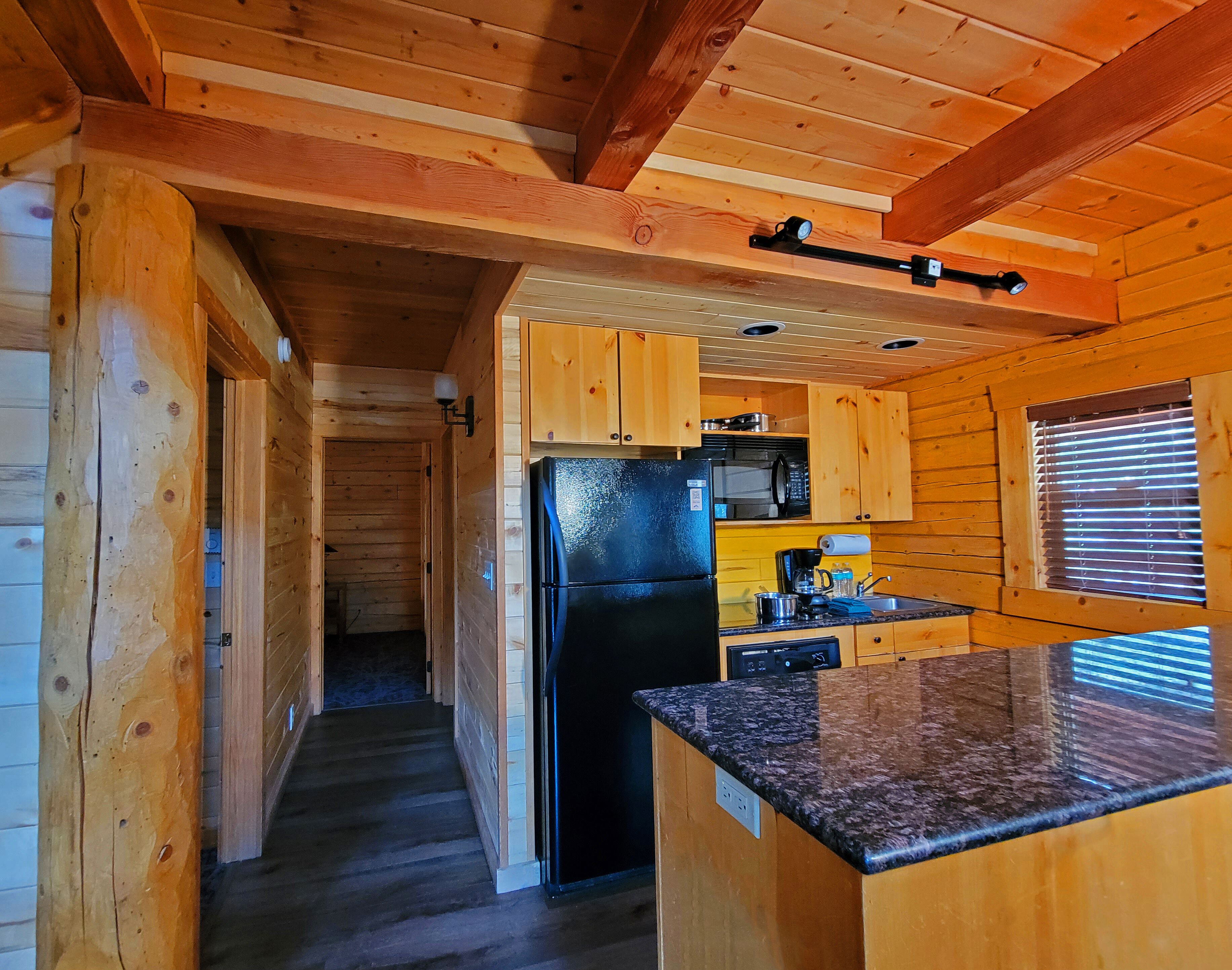 L’intérieur d’un chalet en bois comprend un coin cuisine avec un réfrigérateur noir, des armoires en bois et un plan de travail en granit.