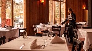 Un restaurante con paredes de madera y ventanas amplias, mesas dispuestas con manteles y servilletas blancas, y un camarero atendiendo una mesa.