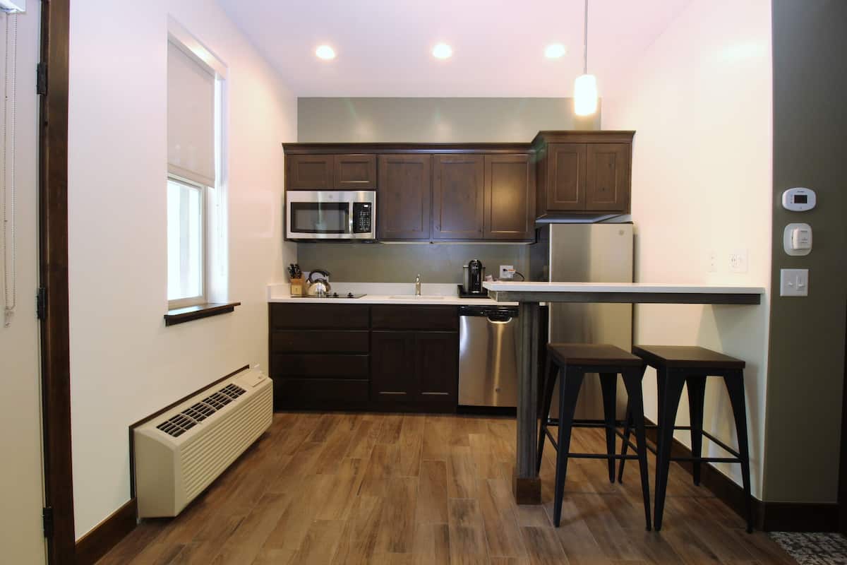 Una cocina compacta con armarios de madera oscura, un microondas de acero inoxidable, lavavajillas y refrigerador. Hay una ventana con persianas, un calentador integrado y un taburete en la barra.