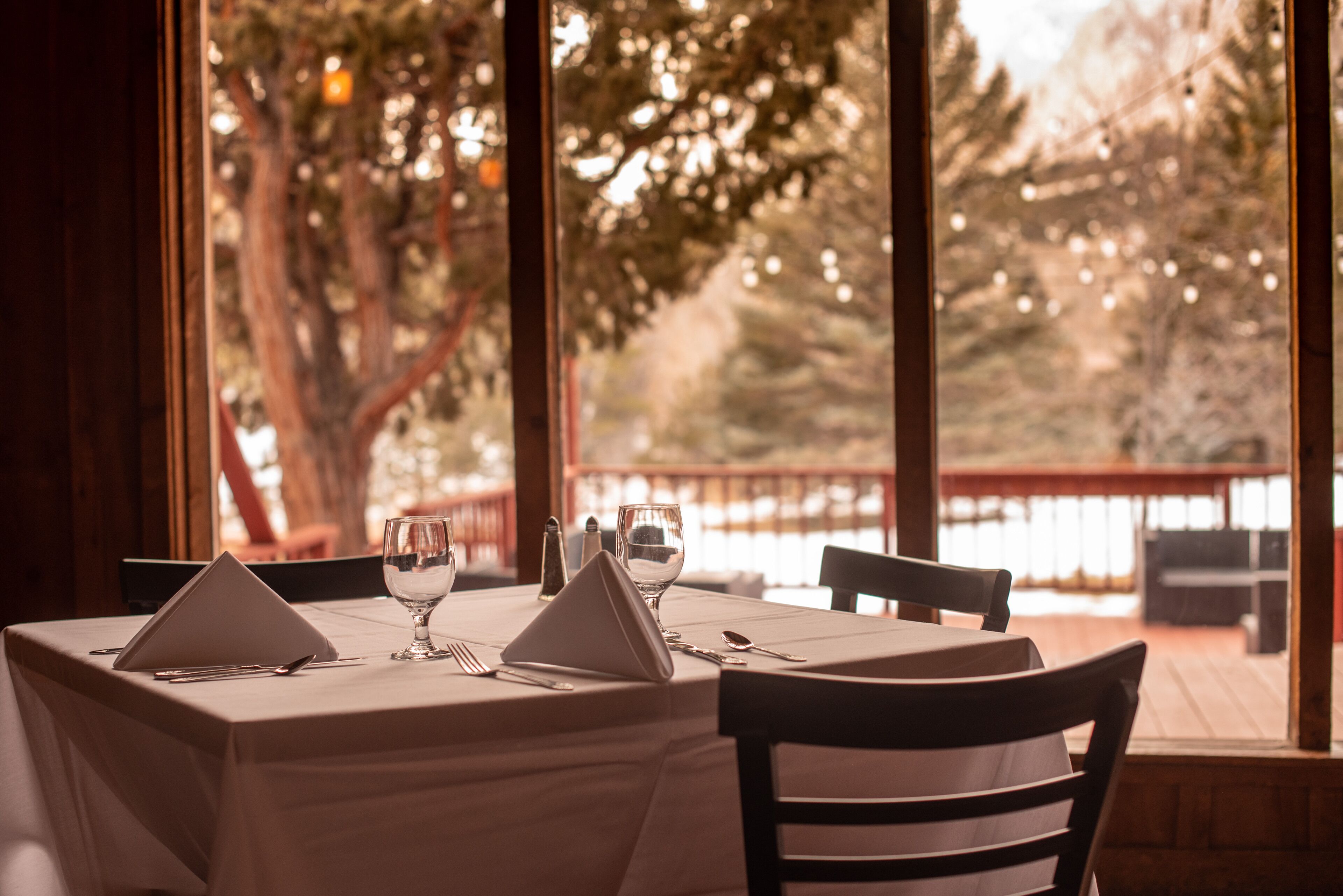 A dining table set for two with a view of trees and a body of water through large windows.