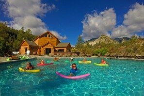 A swimming pool with people on floaties, surrounded by a wooden building and mountains. - Mount Princeton Hot Springs Resort (Nathrop)